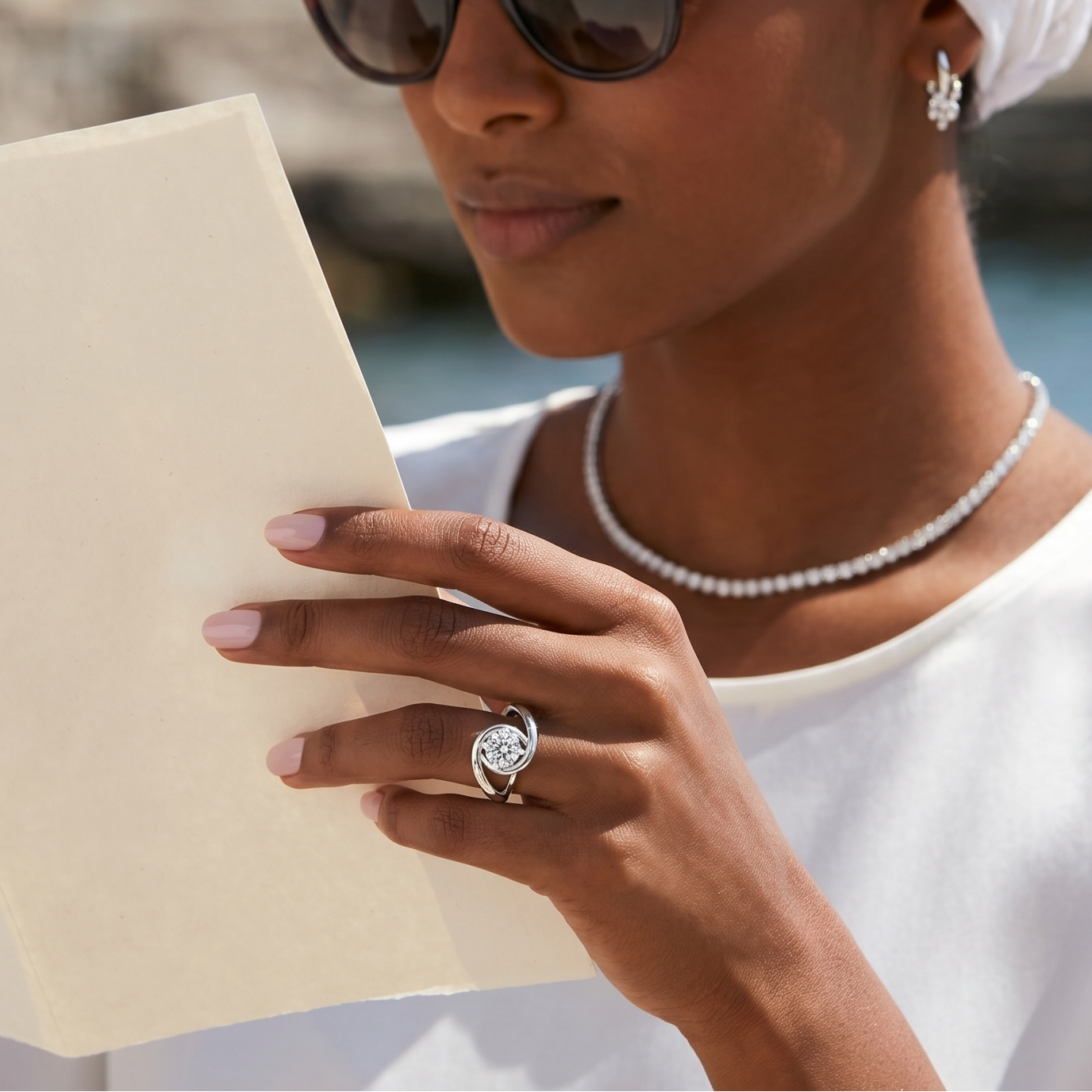 Woman wearing sunglasses and a white headscarf, reading a book by the water.