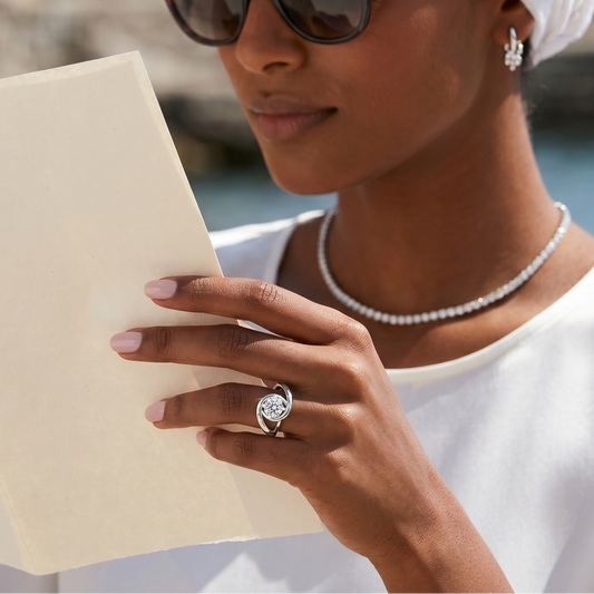 Woman wearing sunglasses and a white headscarf, reading a book by the water.